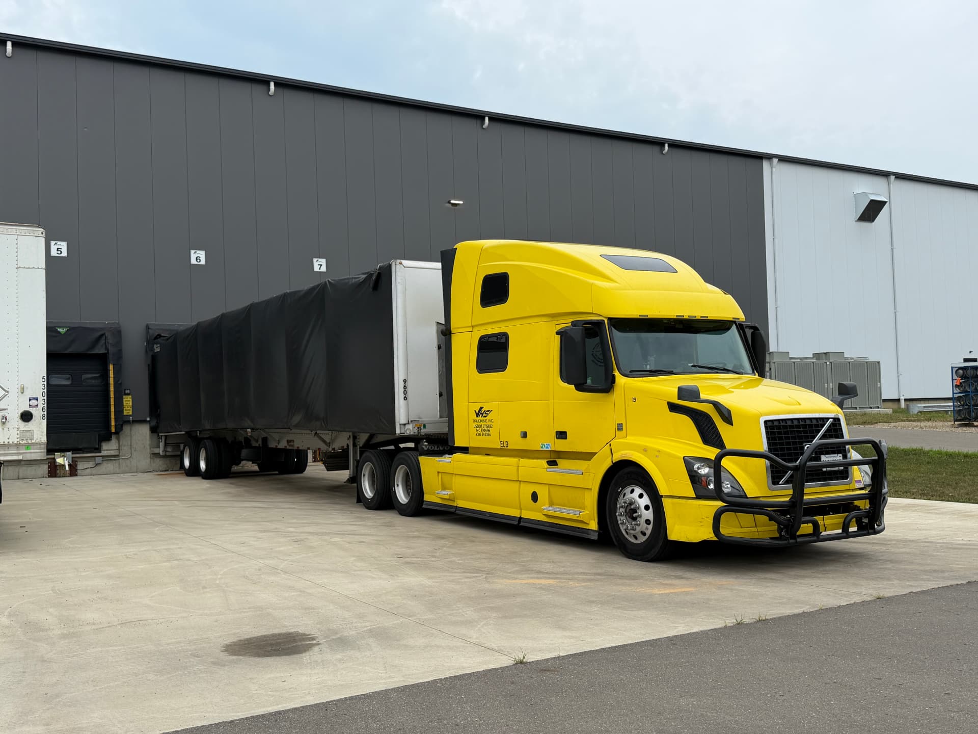 Yellow NK Extra Services truck with a Conestoga trailer loading at a warehouse dock