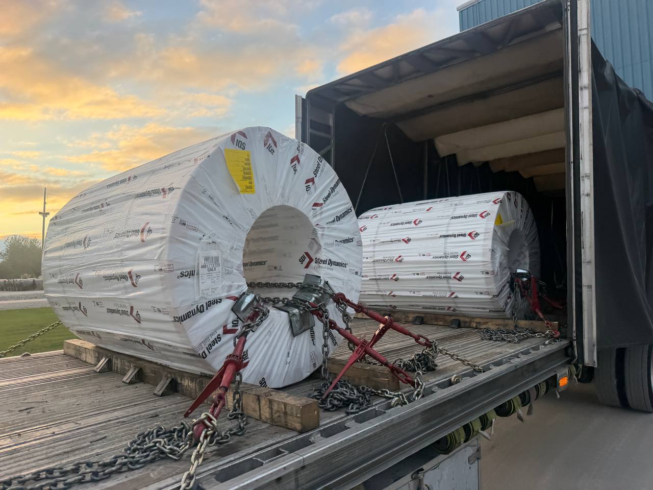 Two wrapped Steel Dynamics coils chained inside a Conestoga trailer at sunset
